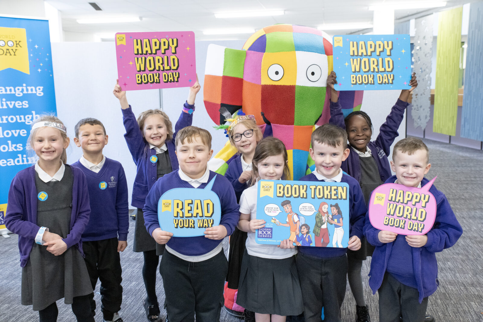 A group of cheerful children dressed in school uniforms celebrate World Book Day, holding colorful signs that read "Happy World Book Day," "Read Your Way," and "Book Token," while standing next to a large, colorful stuffed elephant.
