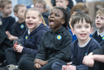 A group of young children sits on the floor, smiling and engaged. The girl in the foreground, wearing a black hoodie, joyfully sings or shouts, while her classmates express excitement. They are wearing matching black and white attire, with colorful badges visible on their clothing.
