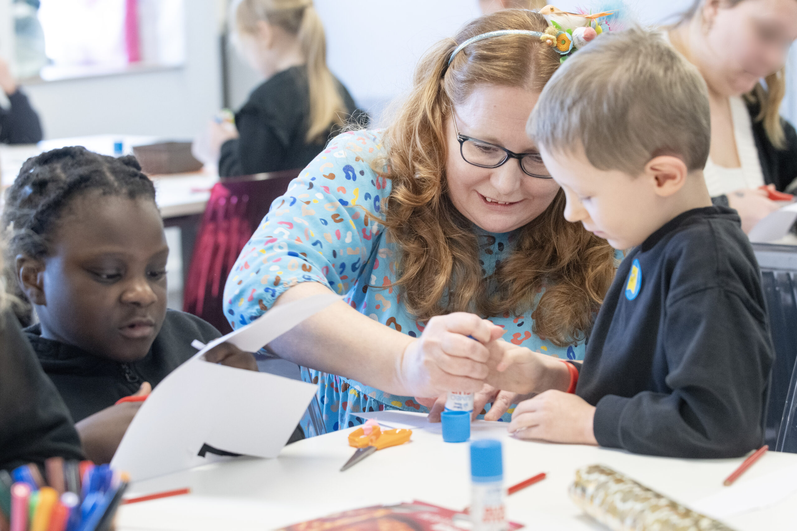 A teacher assists a young boy in using glue on a project, while a girl observes nearby. The scene takes place in a bright classroom filled with art supplies and children engaged in creative activities.