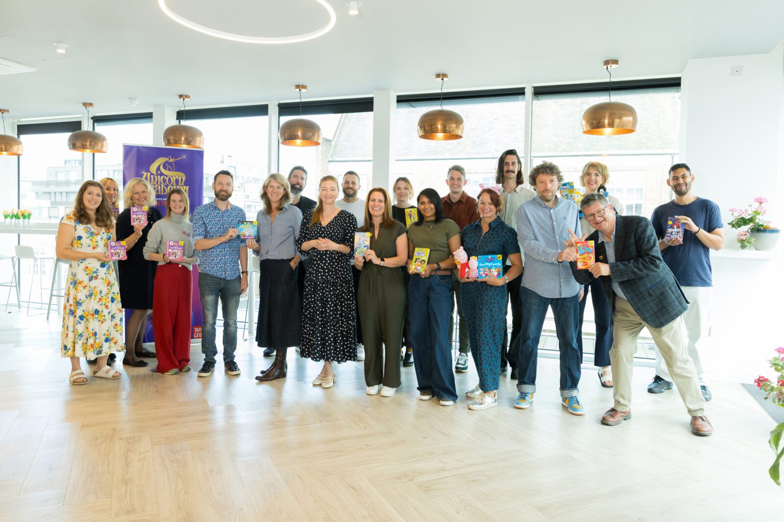 A diverse group of individuals stands together in a bright, modern space, smiling and holding colorful products. The scene conveys a sense of celebration and community among the participants, who are dressed in various styles, showcasing their enthusiasm for the items they represent.