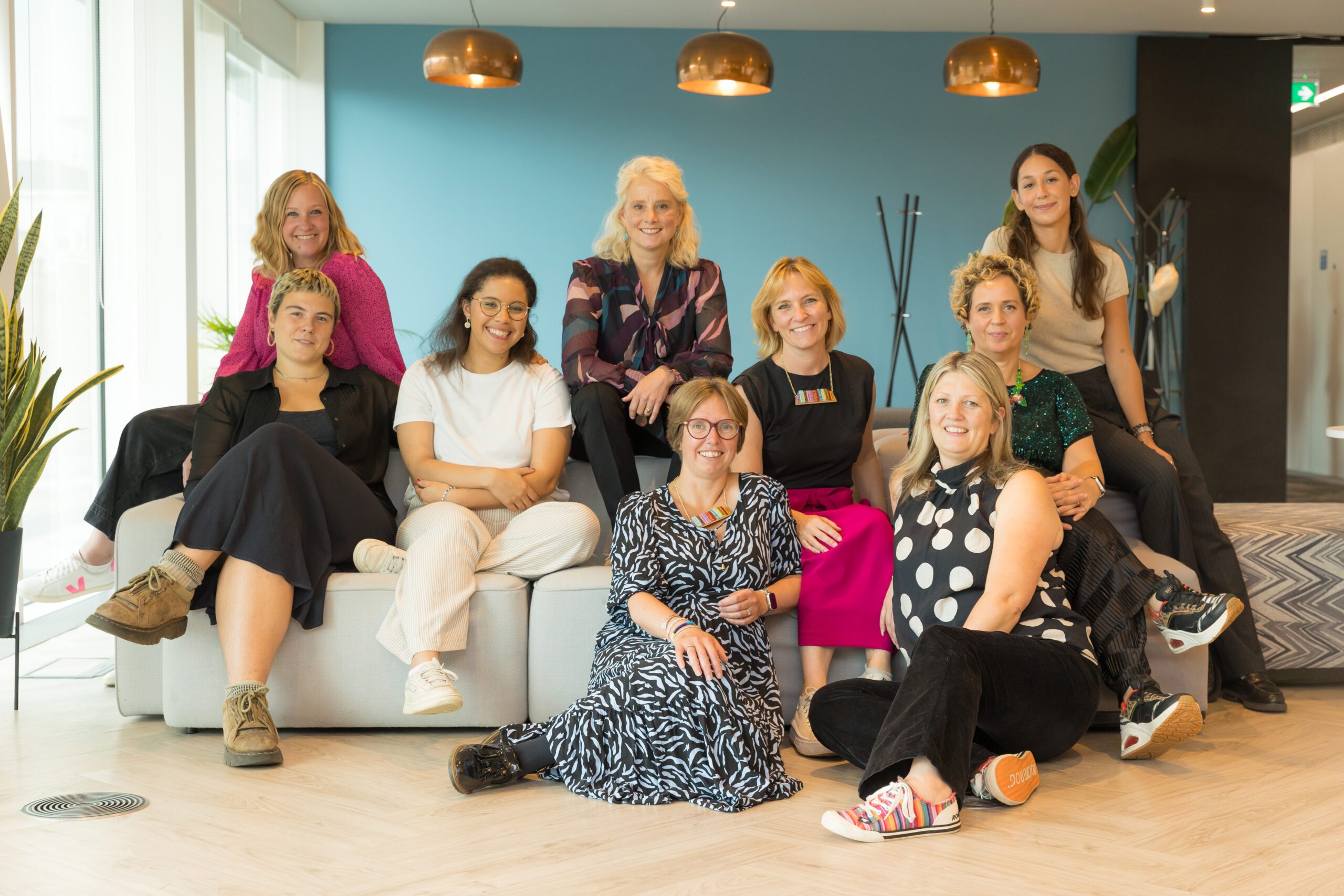 A diverse group of ten women poses together in a modern office space, seated on a sofa. They exhibit a range of clothing styles and hairstyles, smiling and looking at the camera against a backdrop of large windows and stylish decor.