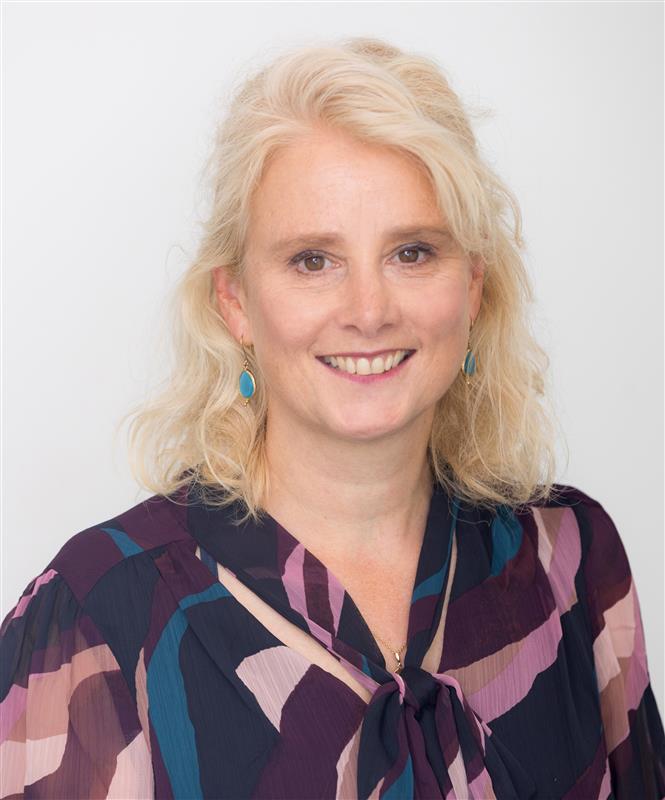 A woman with wavy, blonde hair smiles warmly at the camera. She wears a colorful, patterned blouse and blue earrings, against a plain white background.