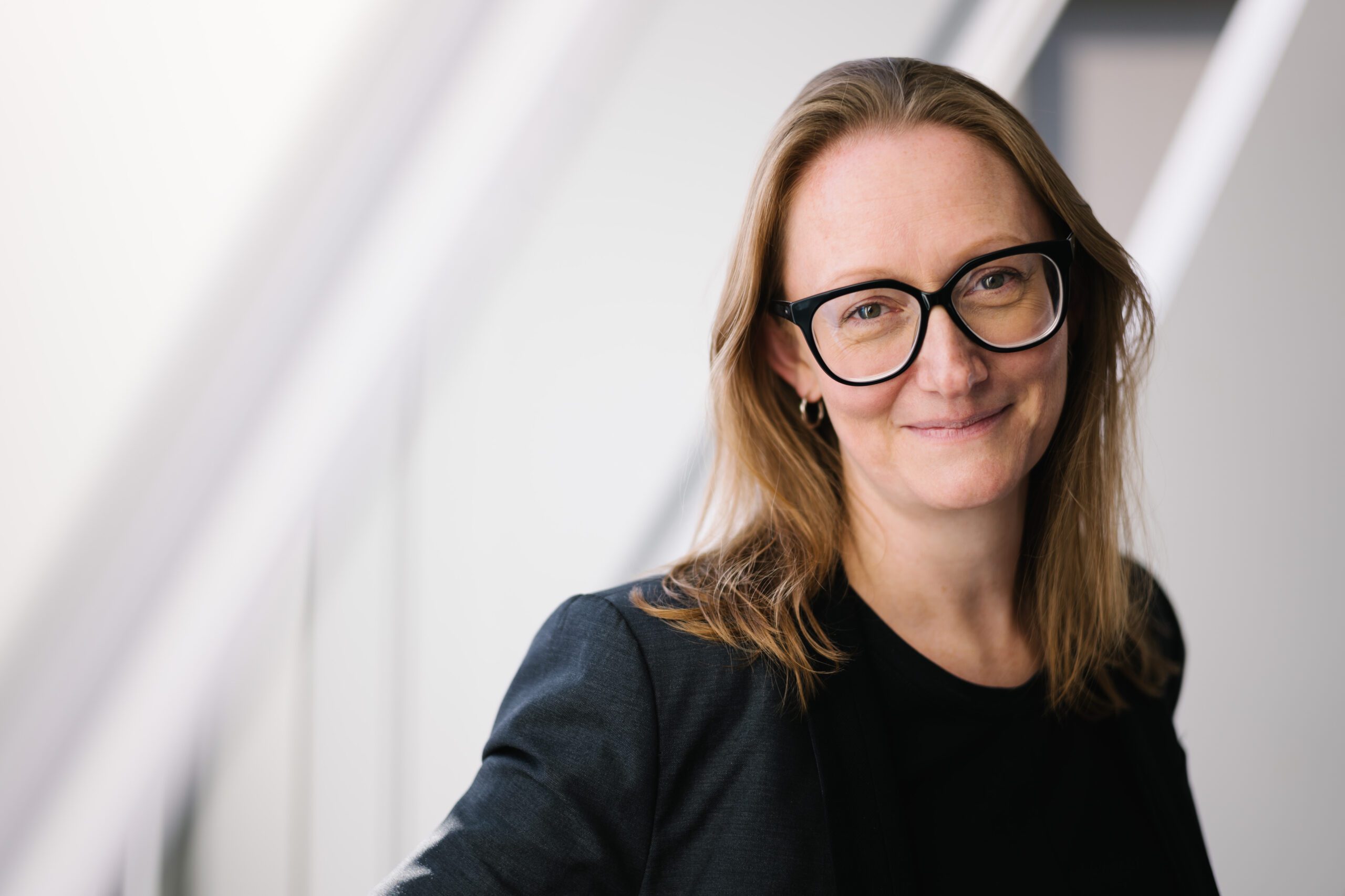 A woman with long hair and glasses, wearing a dark blazer, is smiling softly while leaning against a light-coloured wall, with diagonal lines in the background.