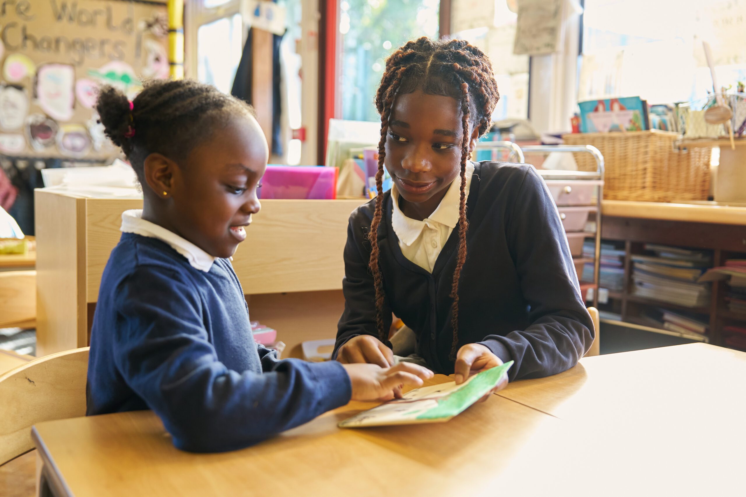 Two schoolchildren in uniforms sit at a wooden table in a classroom, focused on reading a book together.