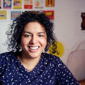 Smiling person with curly hair wearing a patterned navy shirt, seated in front of a colourful wall with artwork.