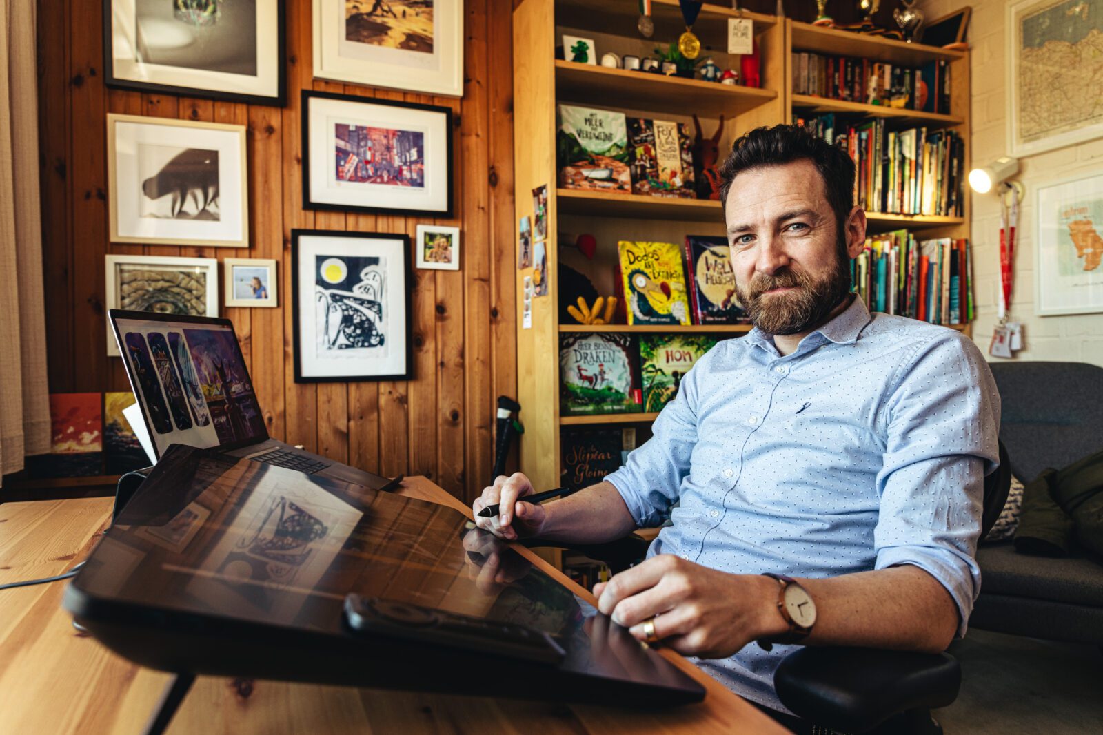 A man sitting at a desk using a digital drawing tablet in a cosy, art-filled office with books and framed pictures on wood-panelled walls.