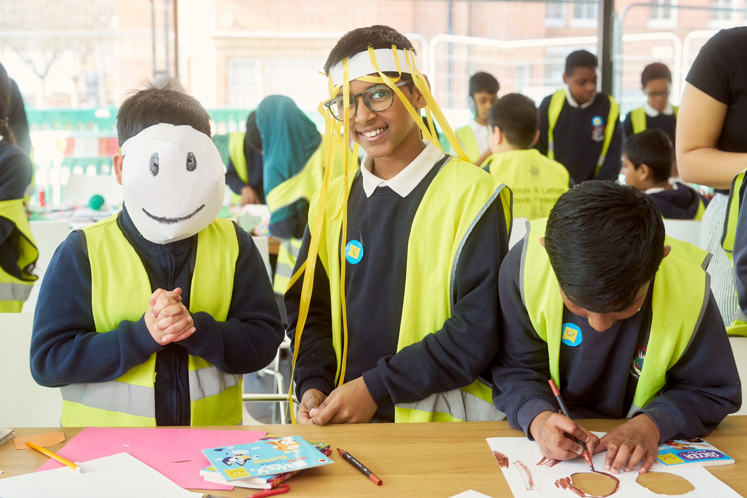 Children wearing hi-vis vests are engaged in a creative activity at a table. One child wears a smiling mask and another has yellow streamers on their head. Others work with paper and pens in the background.