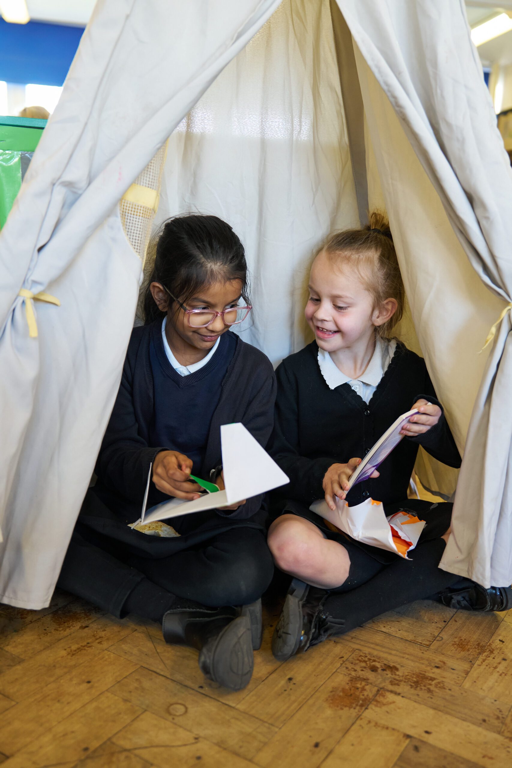 Two schoolgirls sitting inside a fabric tent in a classroom, smiling and engaging in a crafting activity, with paper and scissors.