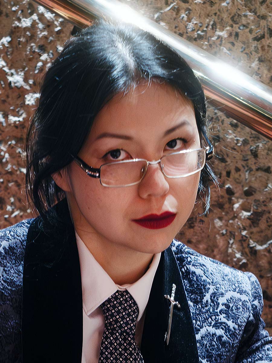 A person with dark hair, red lipstick, and glasses wears a patterned blazer and tie, with a dagger-shaped brooch on the lapel. They are gazing intently at the camera against a textured stone background.