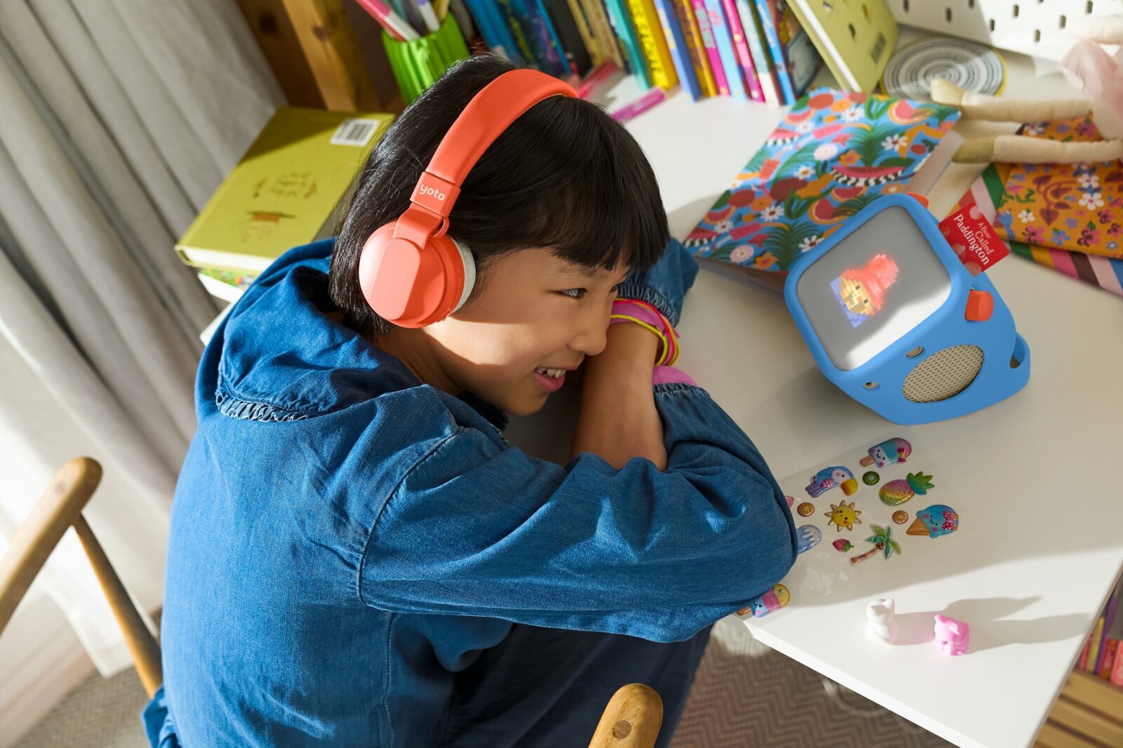 A child wearing orange headphones leans on a desk, smiling at a small blue digital device displaying an animated image. Colourful stickers and books surround the device, creating a playful and creative atmosphere.