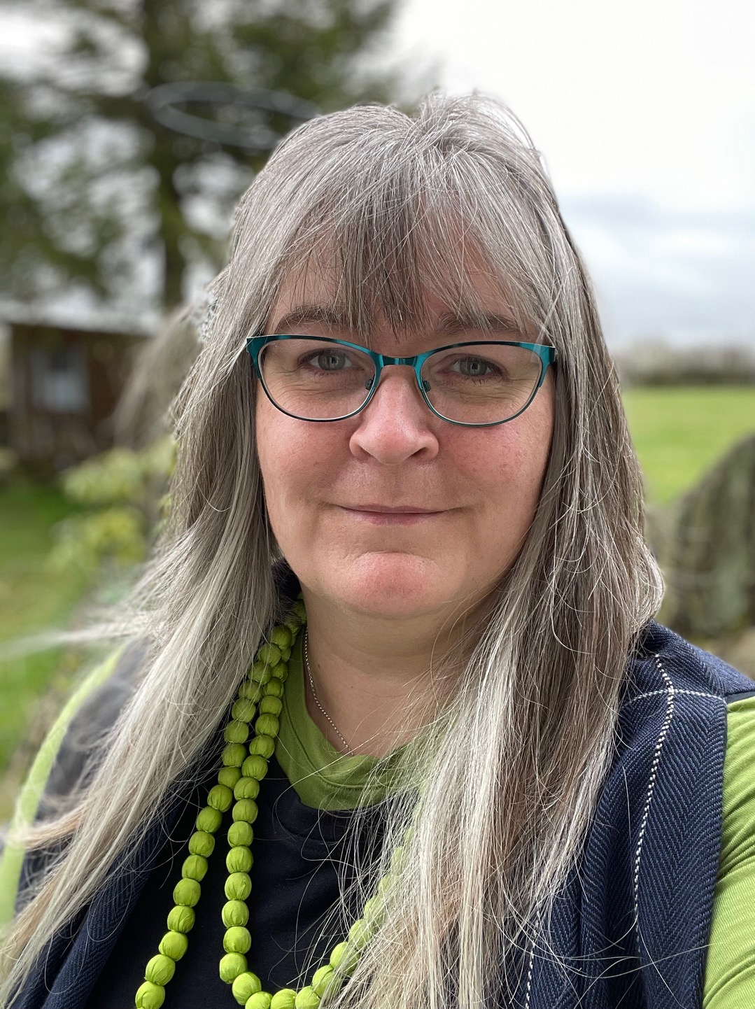 A woman with long grey hair and glasses stands outdoors, wearing a green and black outfit with a beaded necklace, against a blurred background of trees and grassland.