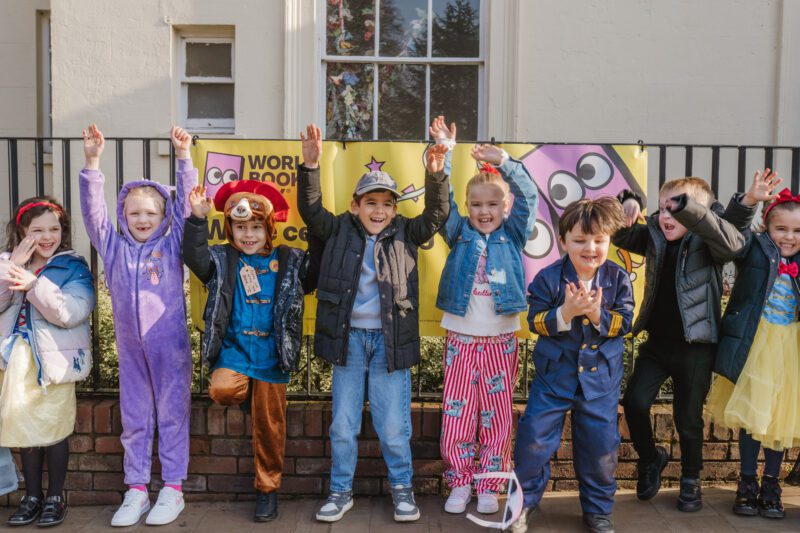 Children in colourful costumes cheerfully pose in front of a yellow banner outdoors.