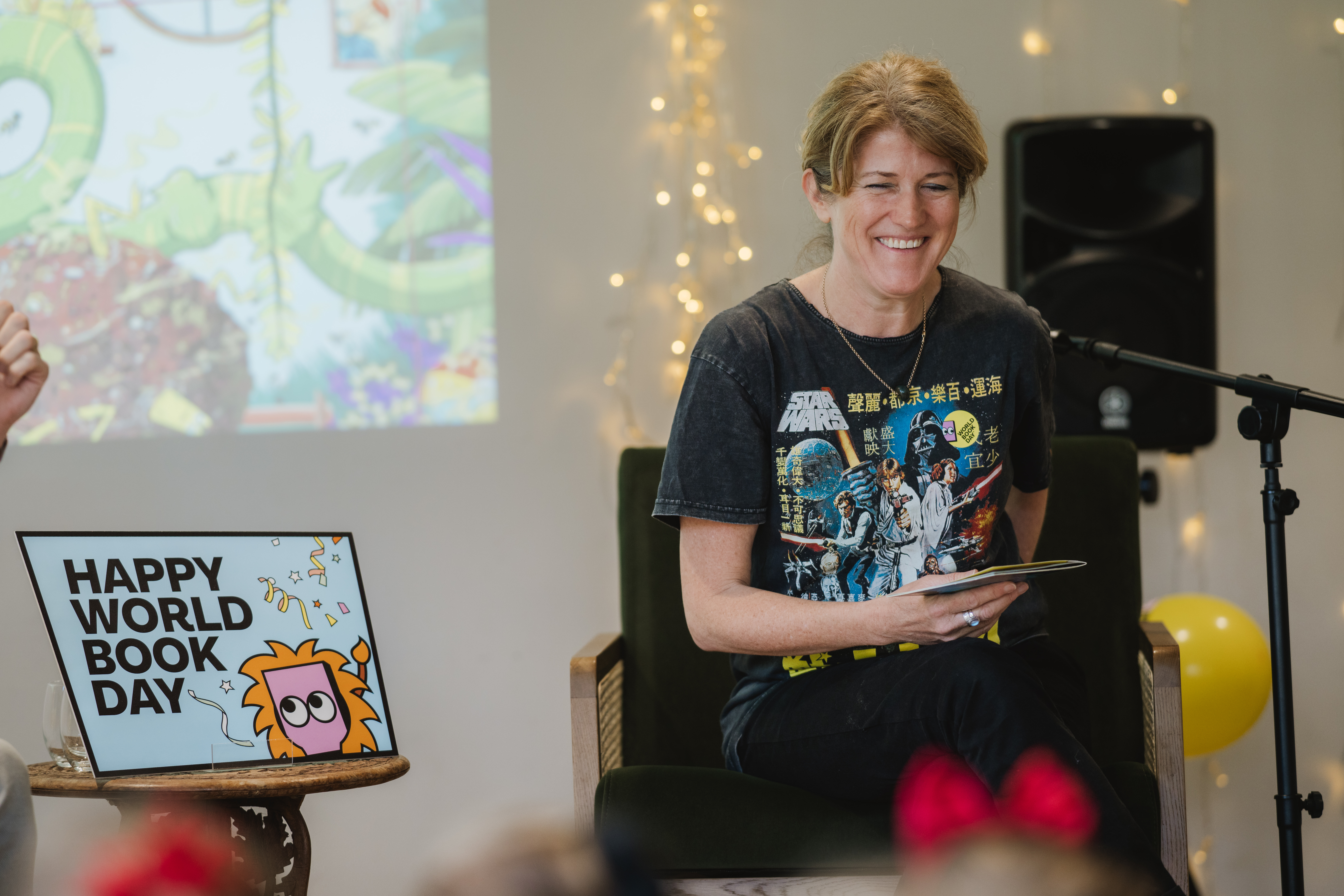 A woman sits on a chair, smiling, while holding a tablet. She's wearing a Star Wars T-shirt. A screen and a "Happy World Book Day" sign are visible, with a background of fairy lights and a speaker.
