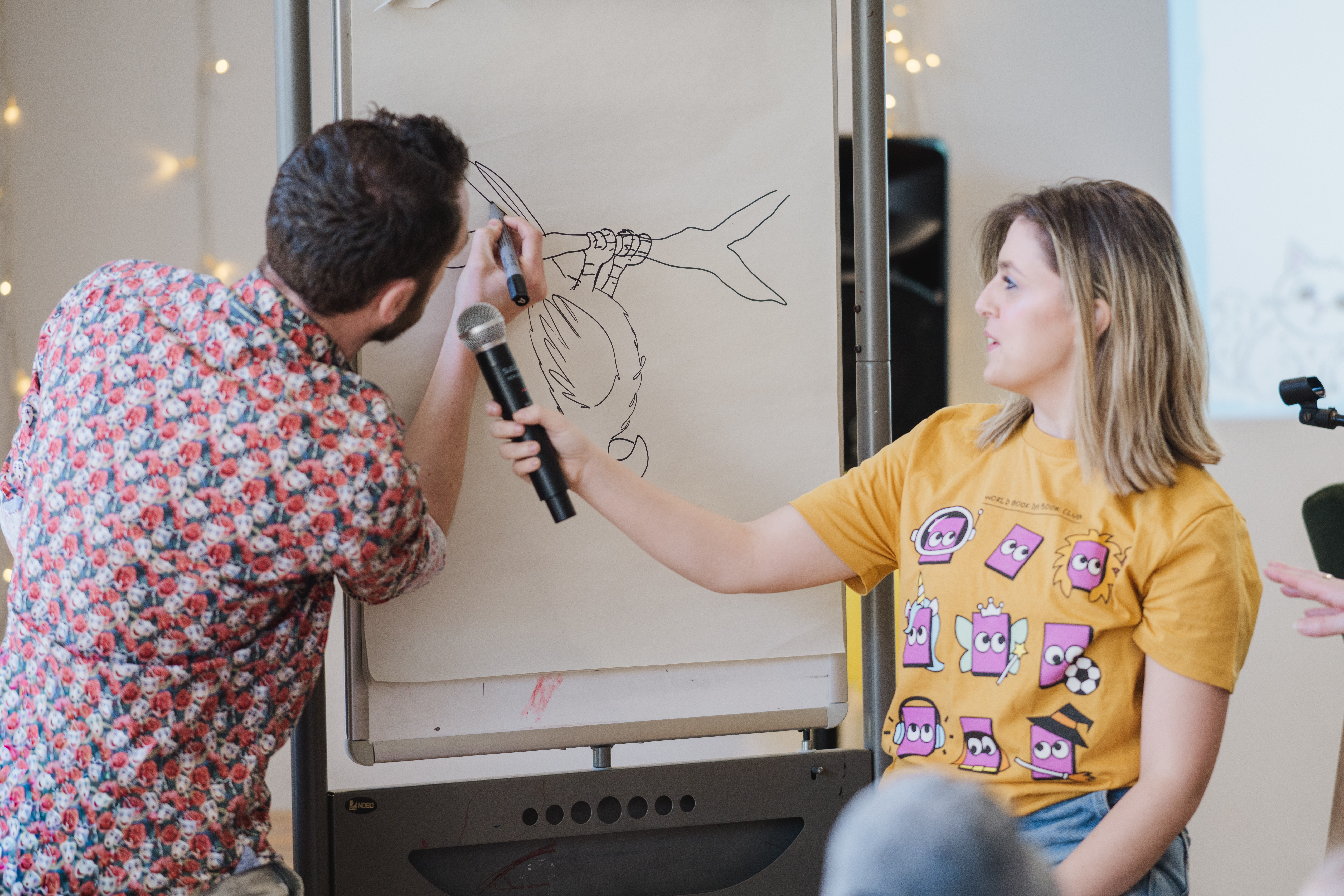 A man draws on a flip chart while a woman holds a microphone for him. She wears a bright yellow T-shirt featuring cartoon faces. The scene appears casual, with both individuals focused on the illustration.