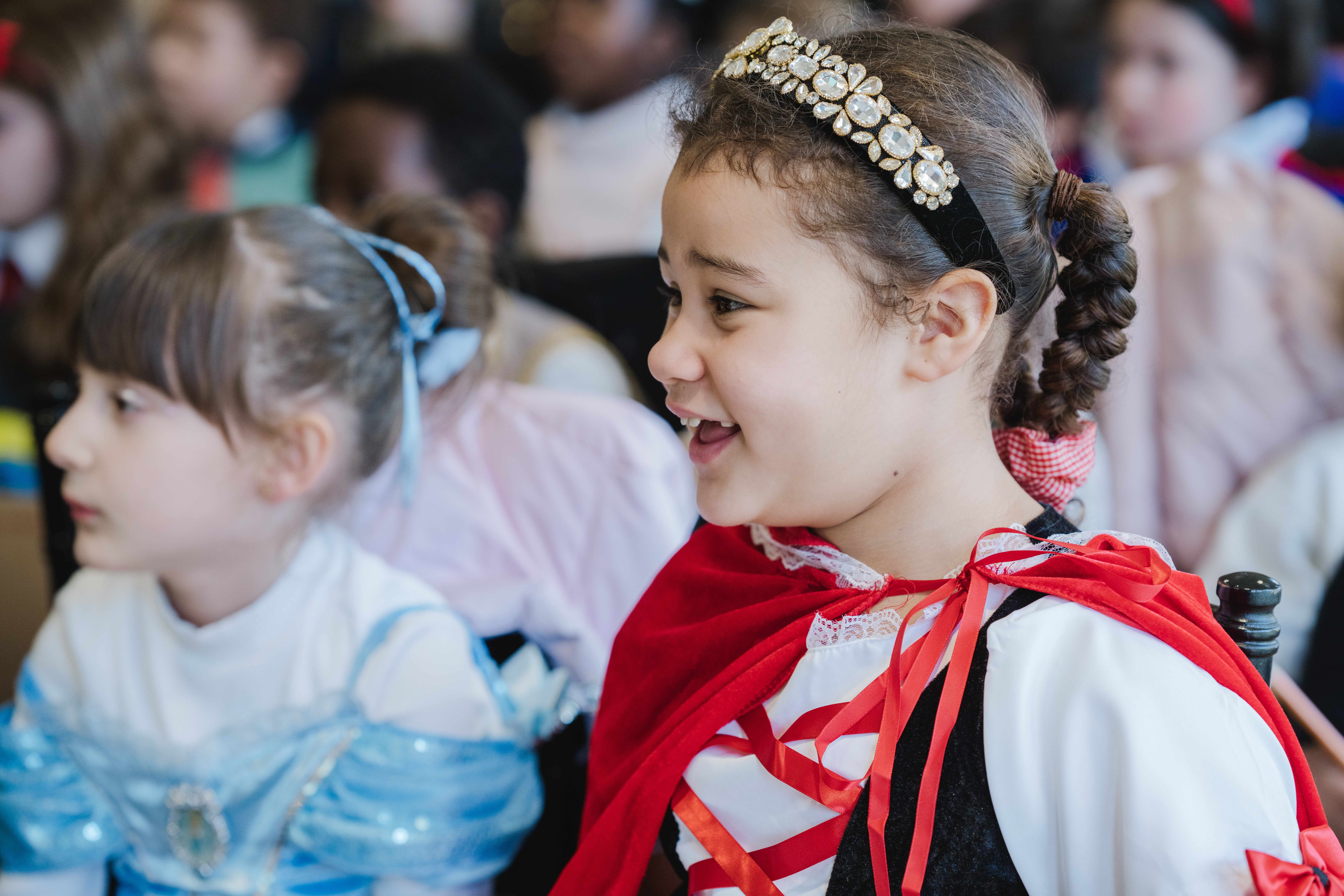 Two young girls in fancy dress costumes, one wearing a tiara, smile and sit among a group of children.