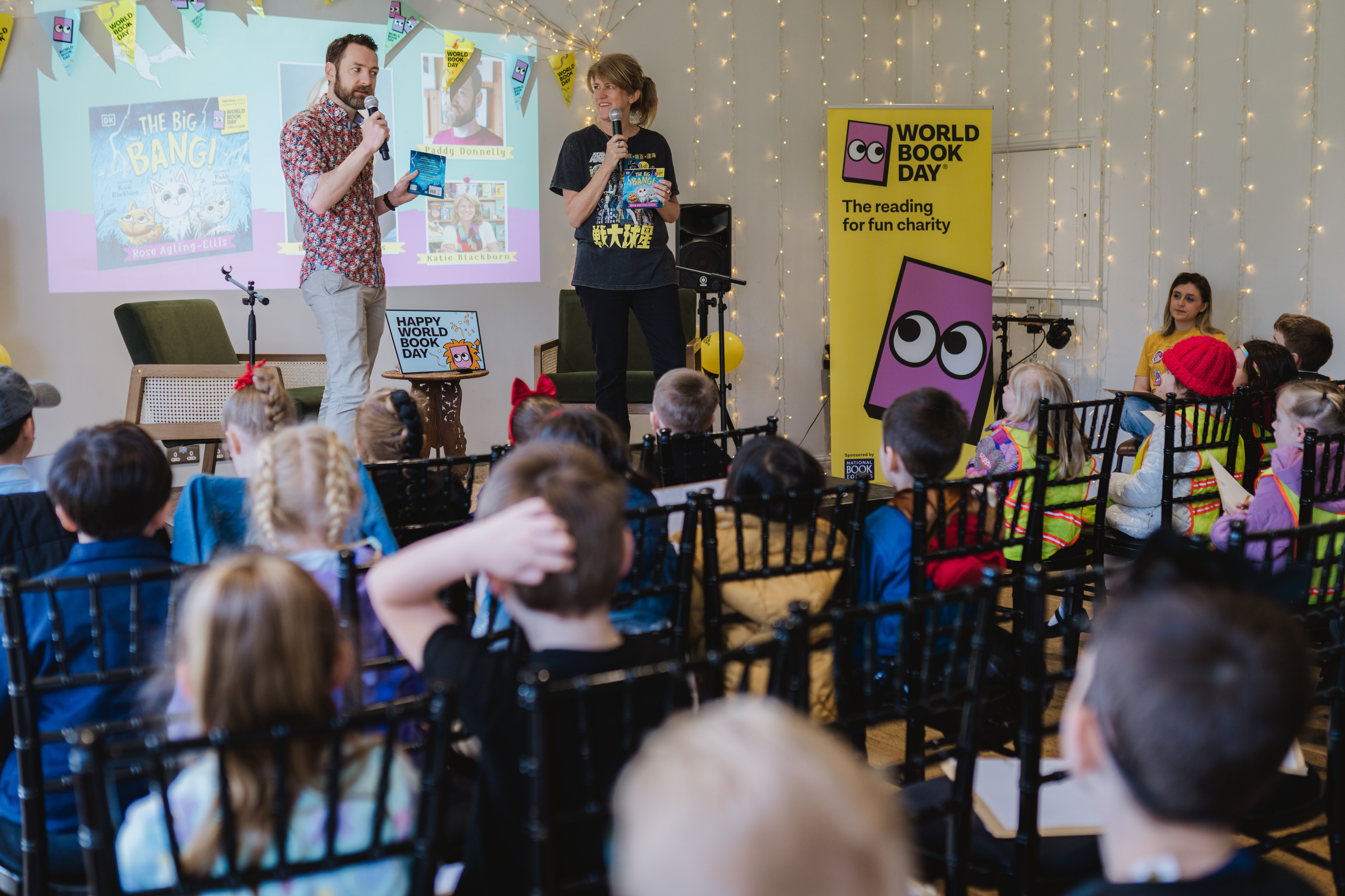 Two presenters stand in front of an audience of children on World Book Day. A yellow banner and book-themed decorations adorn the room.