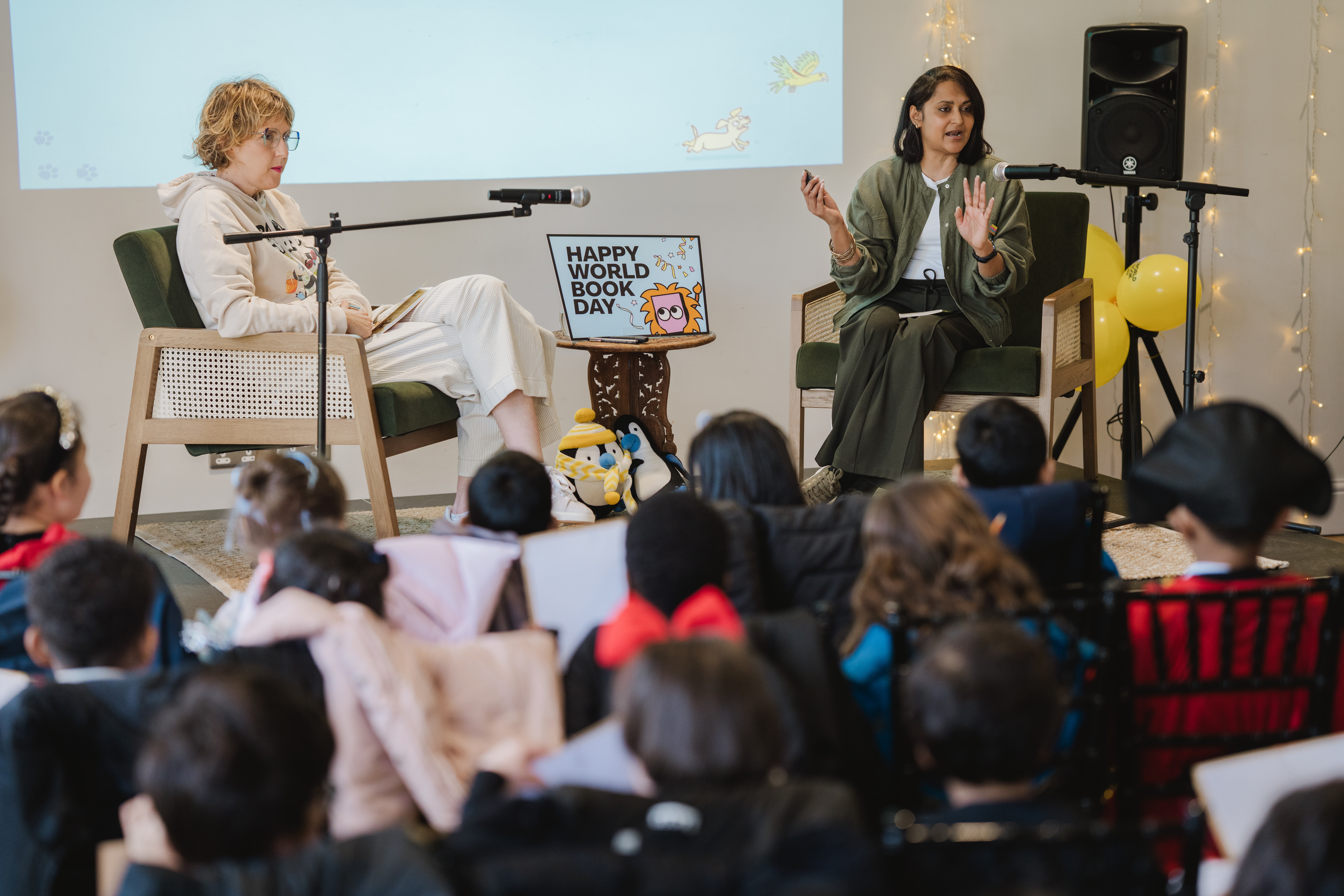 Two people are speaking on stage in front of an audience of children for World Book Day. They are seated in chairs with microphones. A sign reading "Happy World Book Day" is displayed between them. The background includes decorative lights and balloons.