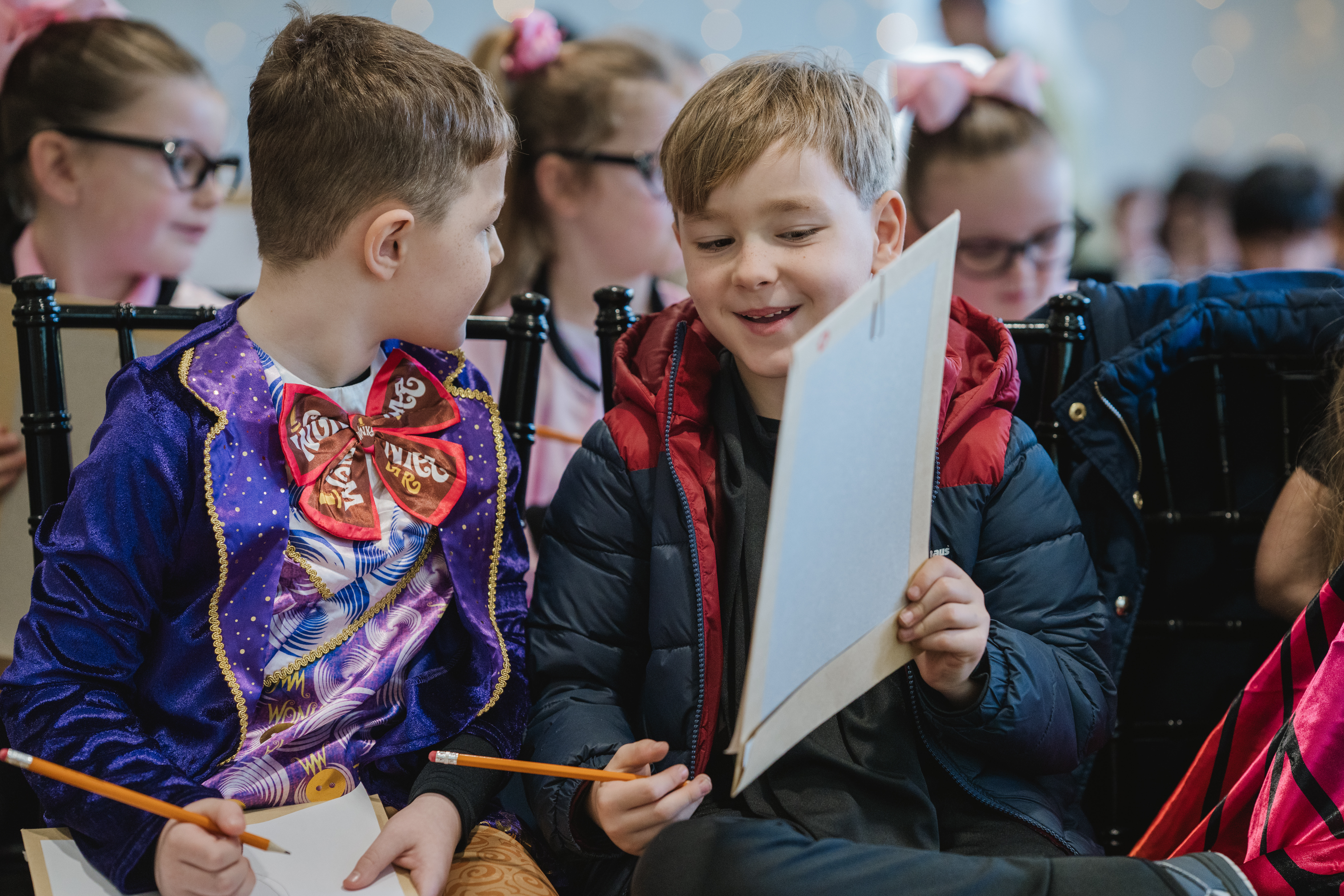 Two young boys in colourful clothing engage in a lively conversation while seated among other children, holding pencils and sketchpads, suggesting a creative activity or workshop setting.
