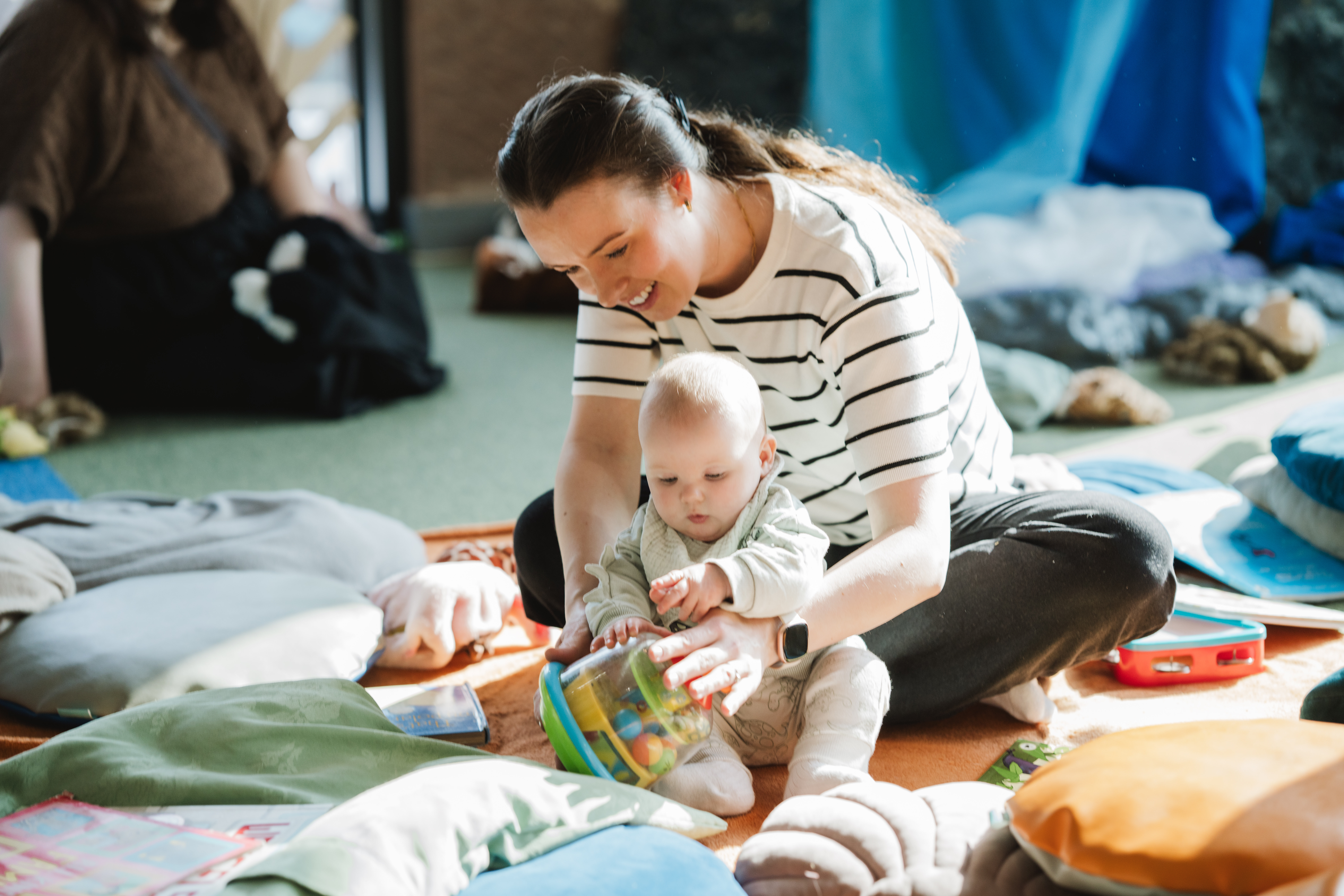 A mother and baby sit on the floor surrounded by cushions and toys. The mother helps the baby play with a colourful toy, both smiling and engaged in a cosy, sunlit room.