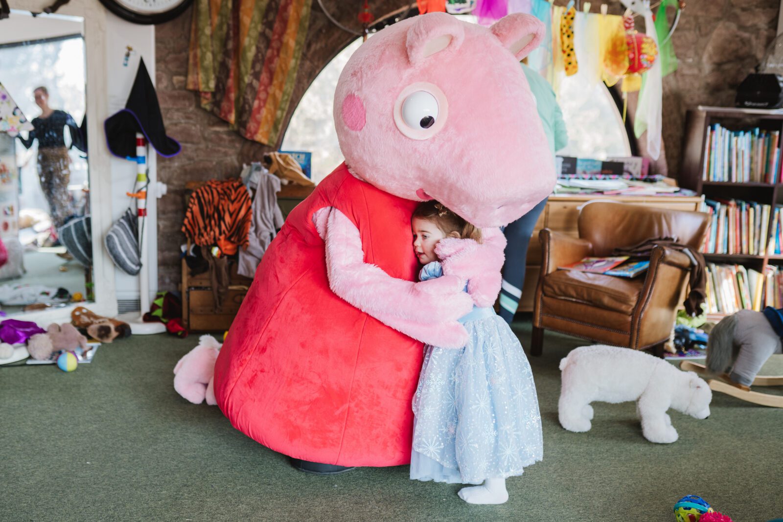 A child in a blue dress hugs a person in a large pink pig costume in a colourful, cluttered room with toys and books.