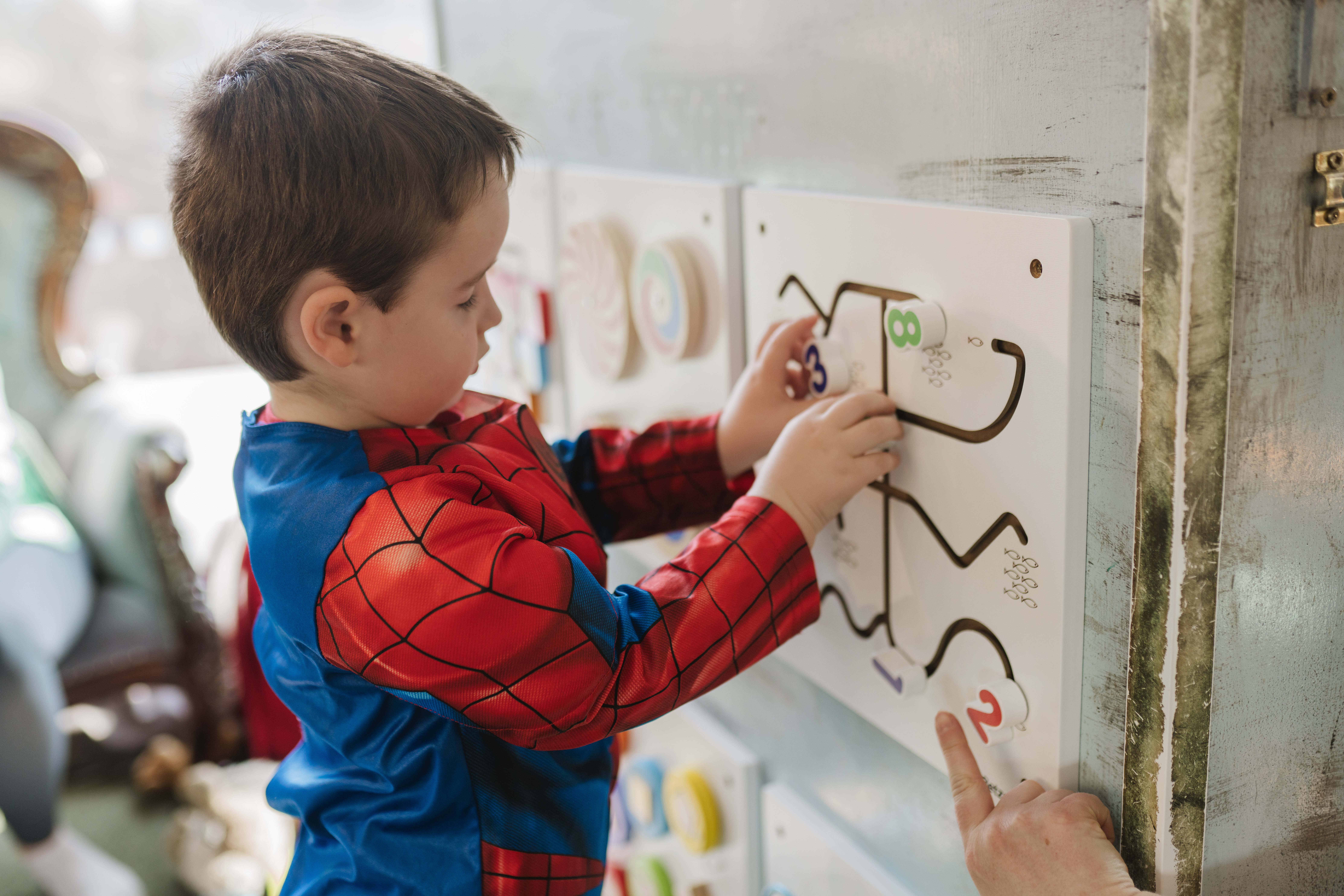 A young boy wearing a Spider-Man costume is playing with an educational wall activity featuring numbers and paths, concentrating as he moves number pieces along the maze.