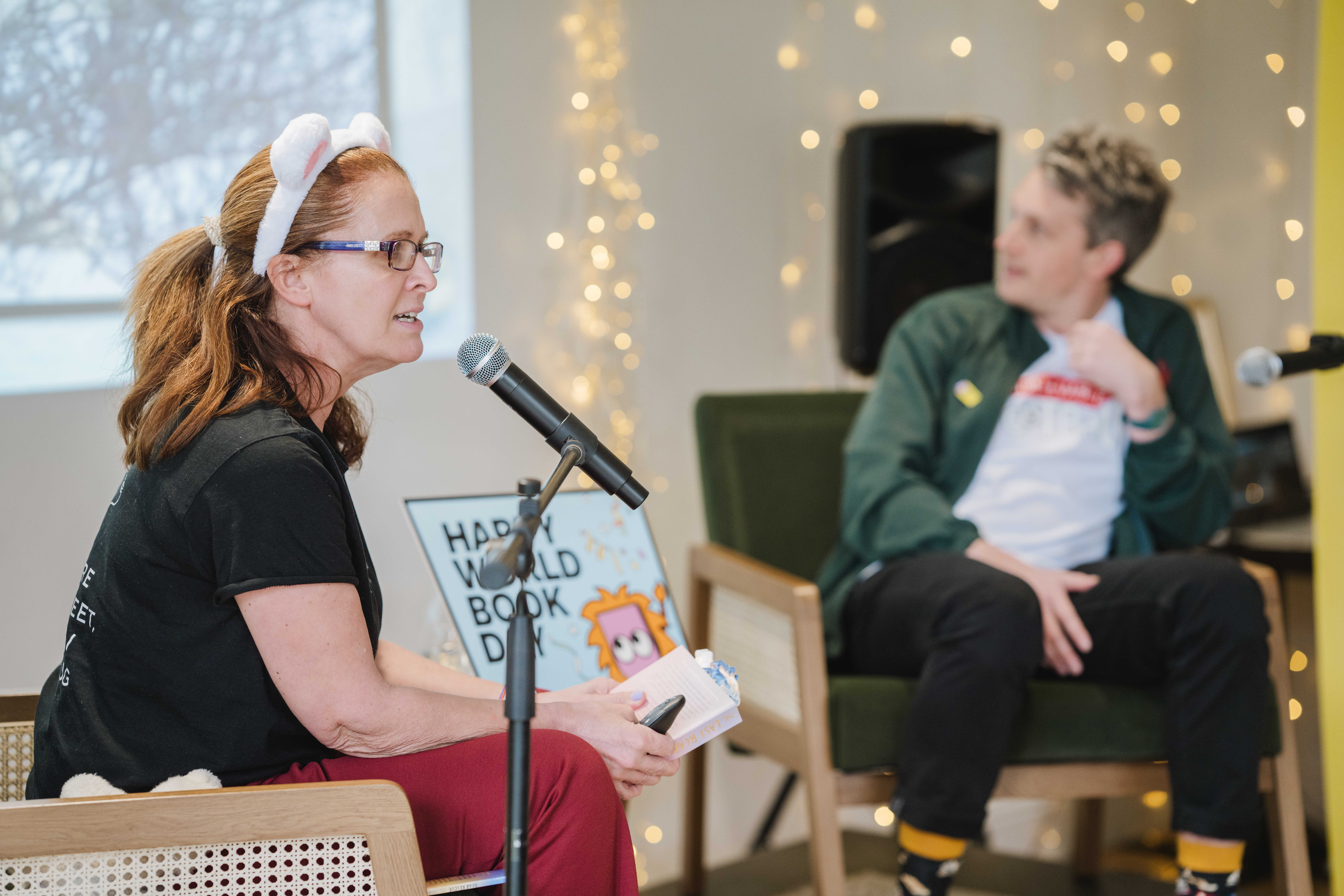 Two people are speaking into microphones in a casually decorated room, with fairy lights in the background. A sign reading "Happy World Book Day" is visible.