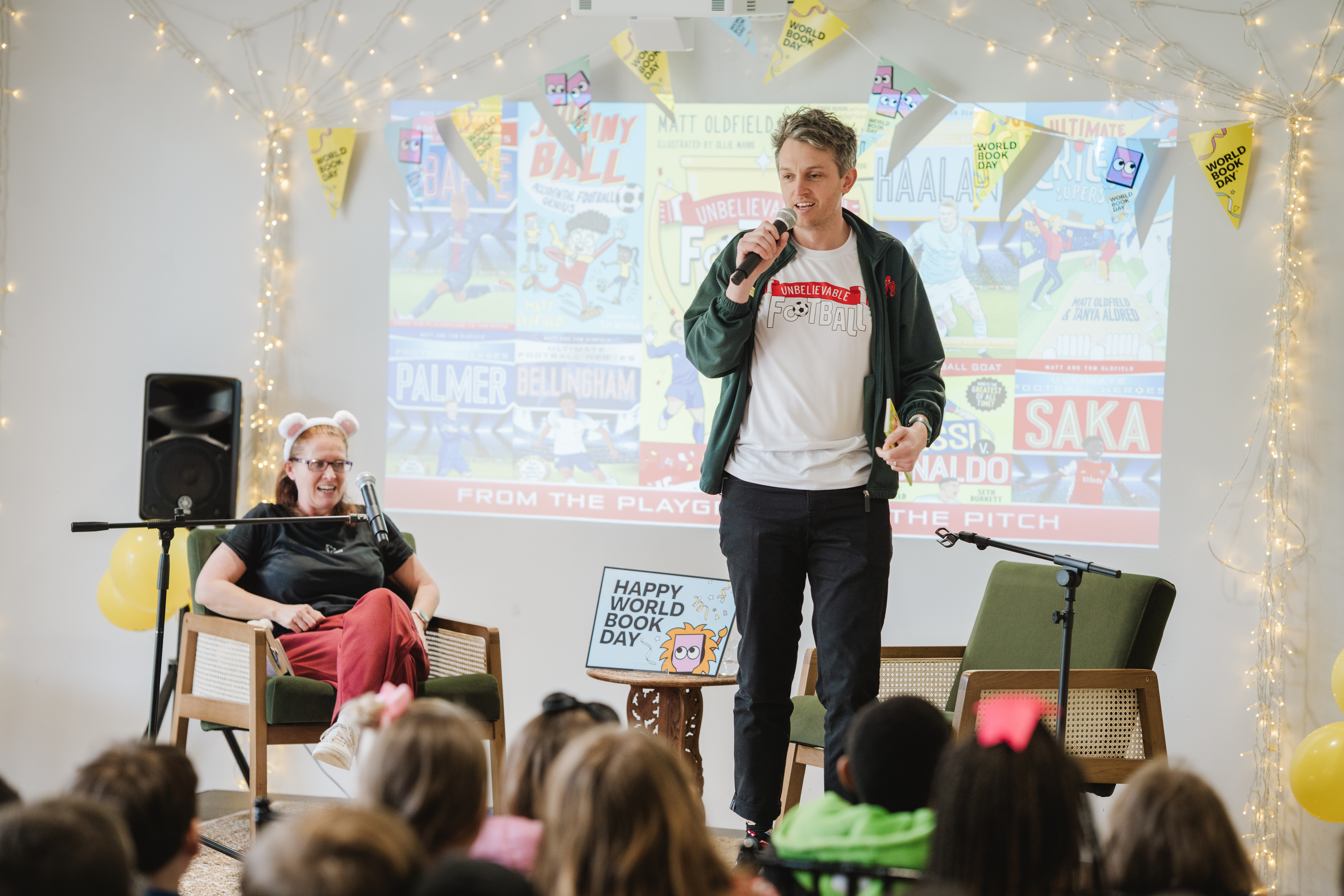 Two presenters engage with children at a World Book Day event, featuring a football theme. One is seated with bear ears, while the other stands with a microphone. A "Happy World Book Day" sign and a football-themed backdrop are visible.