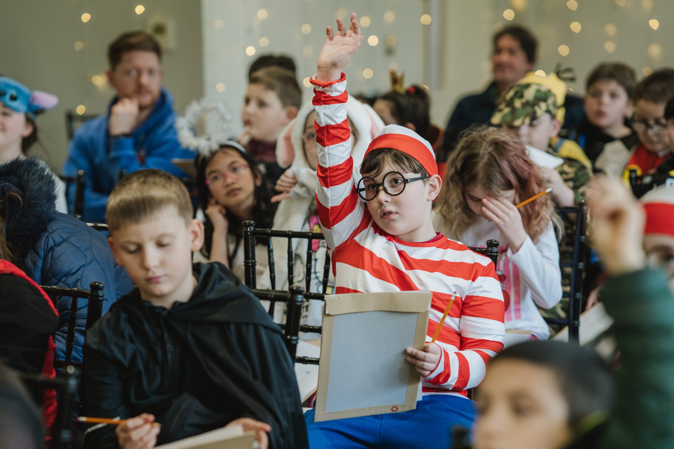 Children in a classroom wearing colourful costumes, including a child dressed in a striped red and white outfit raising a hand, surrounded by peers.