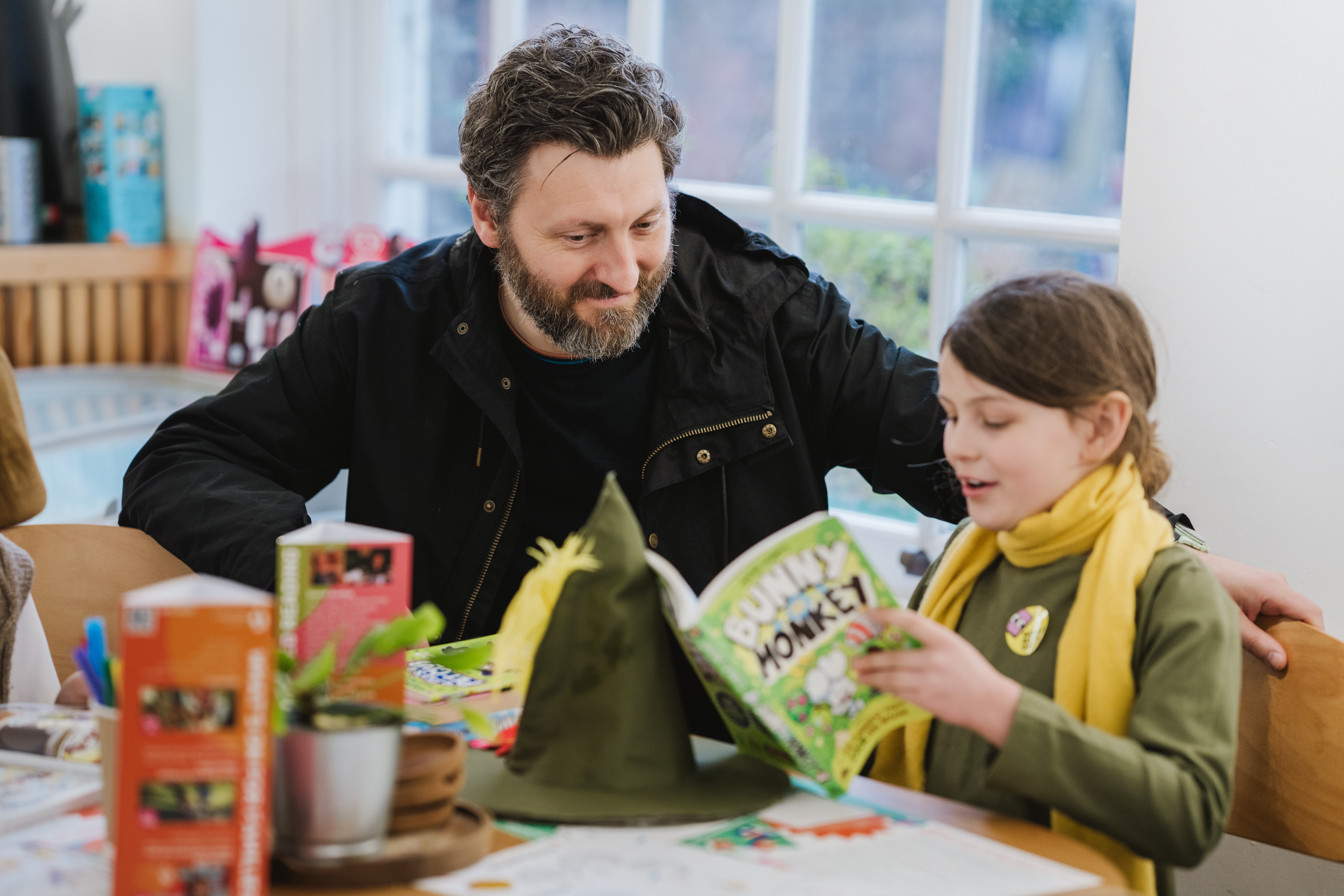 A man and a child are sitting at a table with books and crafts. The child is reading a comic book called "Bunny vs Monkey" aloud, while the man listens attentively. The room has a bright and cosy atmosphere.