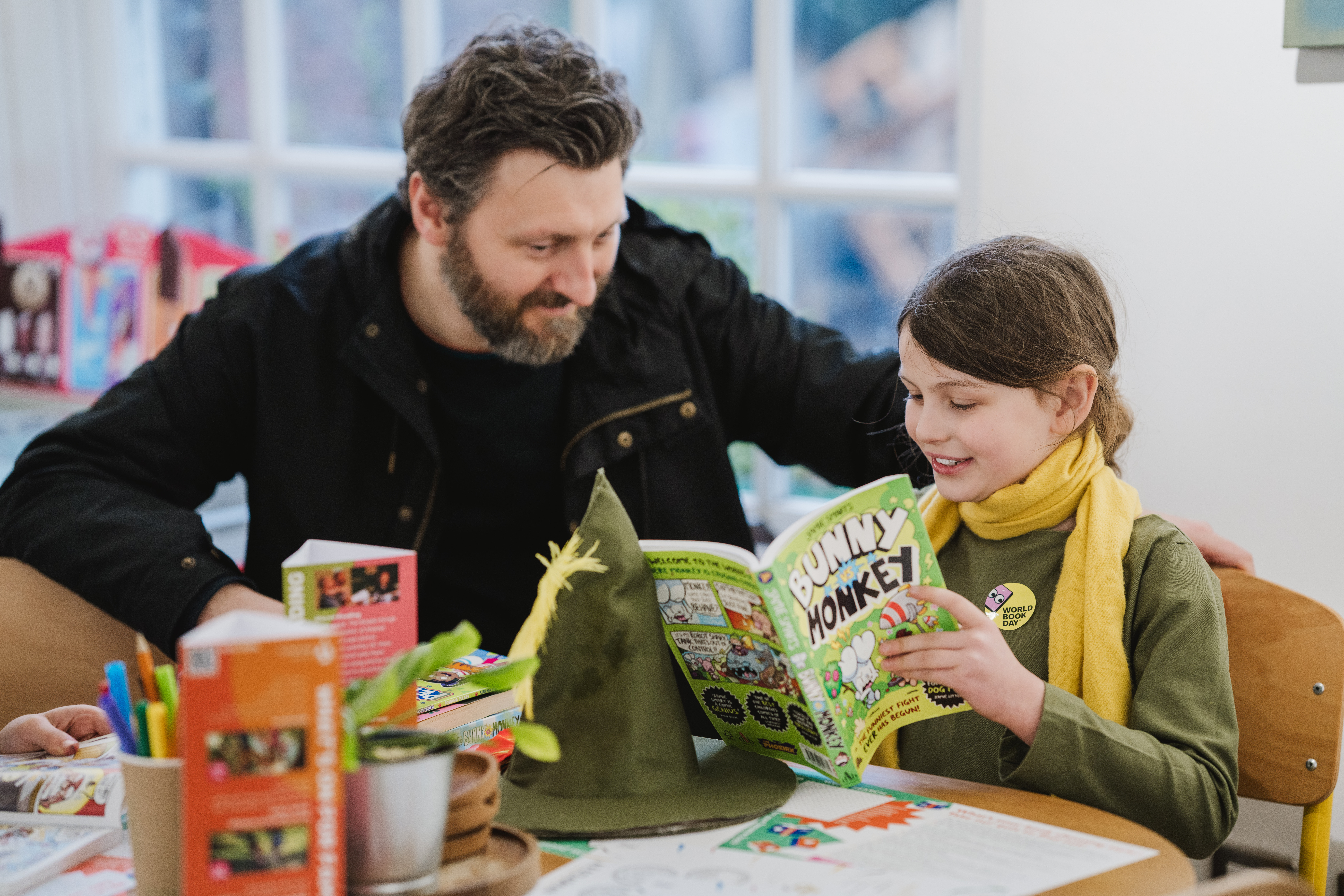 A man and a child sit at a table, reading a "Bunny vs Monkey" comic. The child wears a yellow scarf and both seem engaged. The table has books, coloured pencils, and a green hat.
