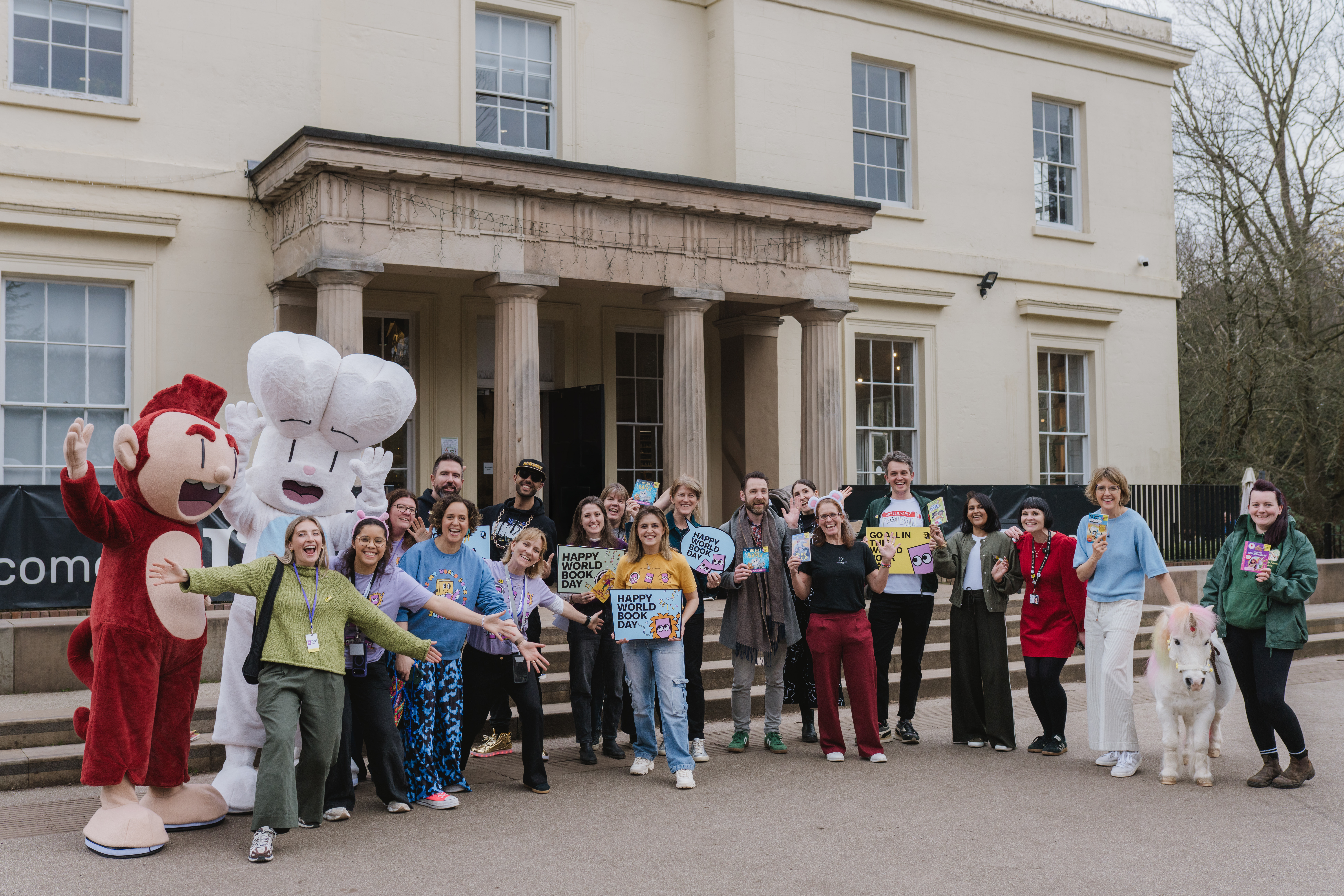 A group of people, including two costume characters and a large fluffy dog, stand cheerfully in front of a grand building. They hold signs celebrating World Book Day, dressed in a variety of casual and themed outfits, conveying a festive atmosphere.