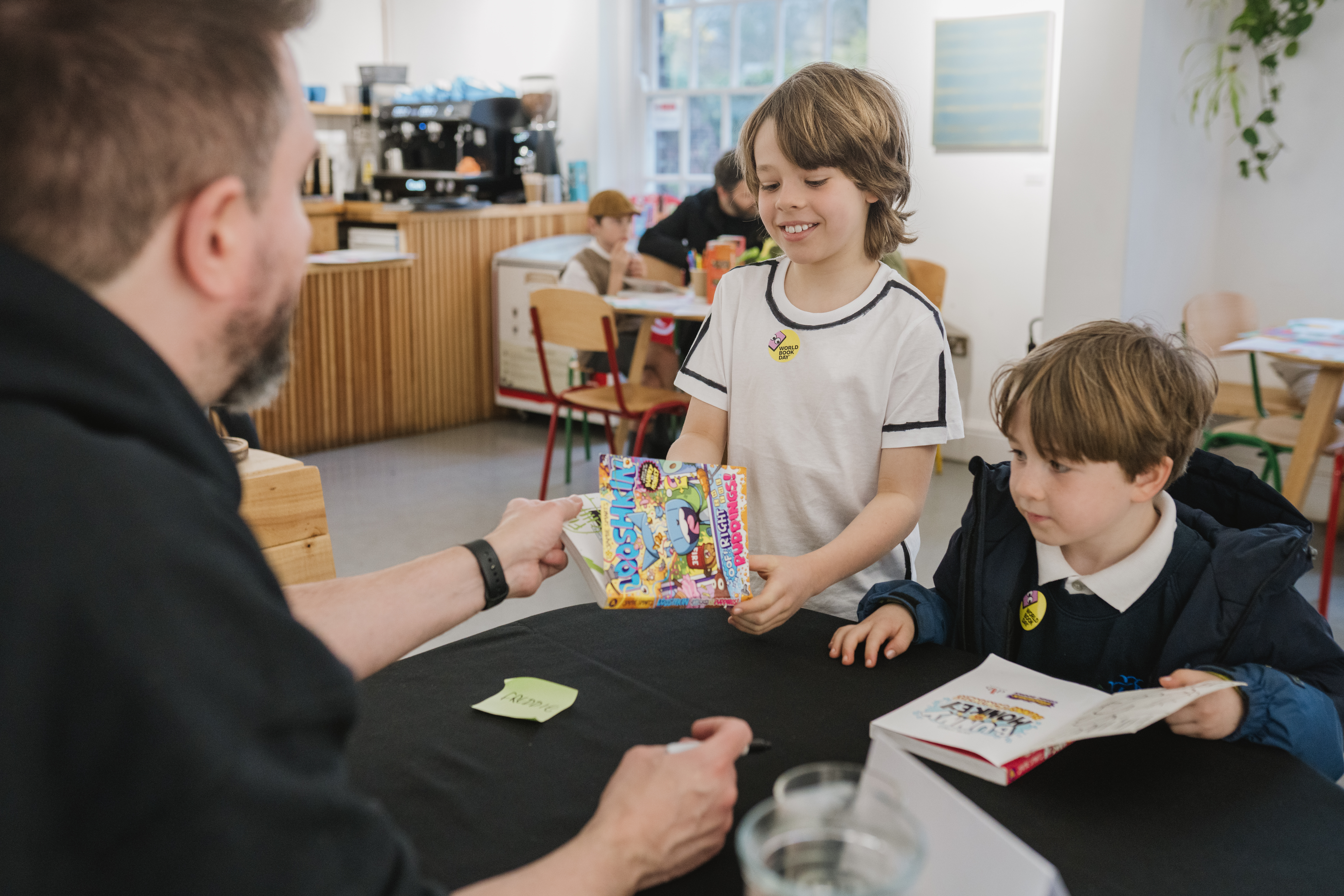 Two young boys at a bookstore counter; one smiling, handing over a colourful book to a seated man.