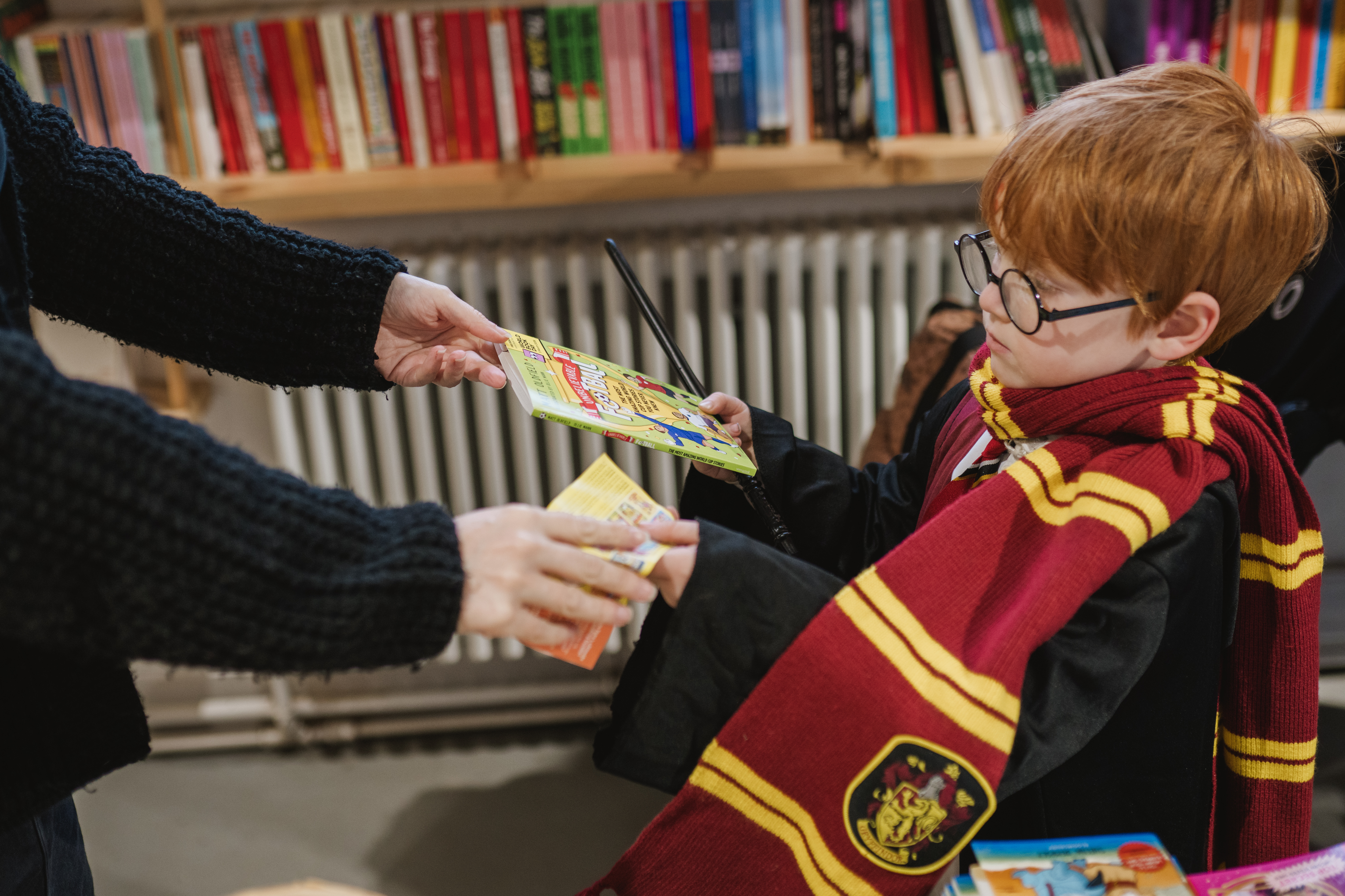 A child dressed as a young wizard wearing glasses and a red and gold scarf receives books from an adult in a library.