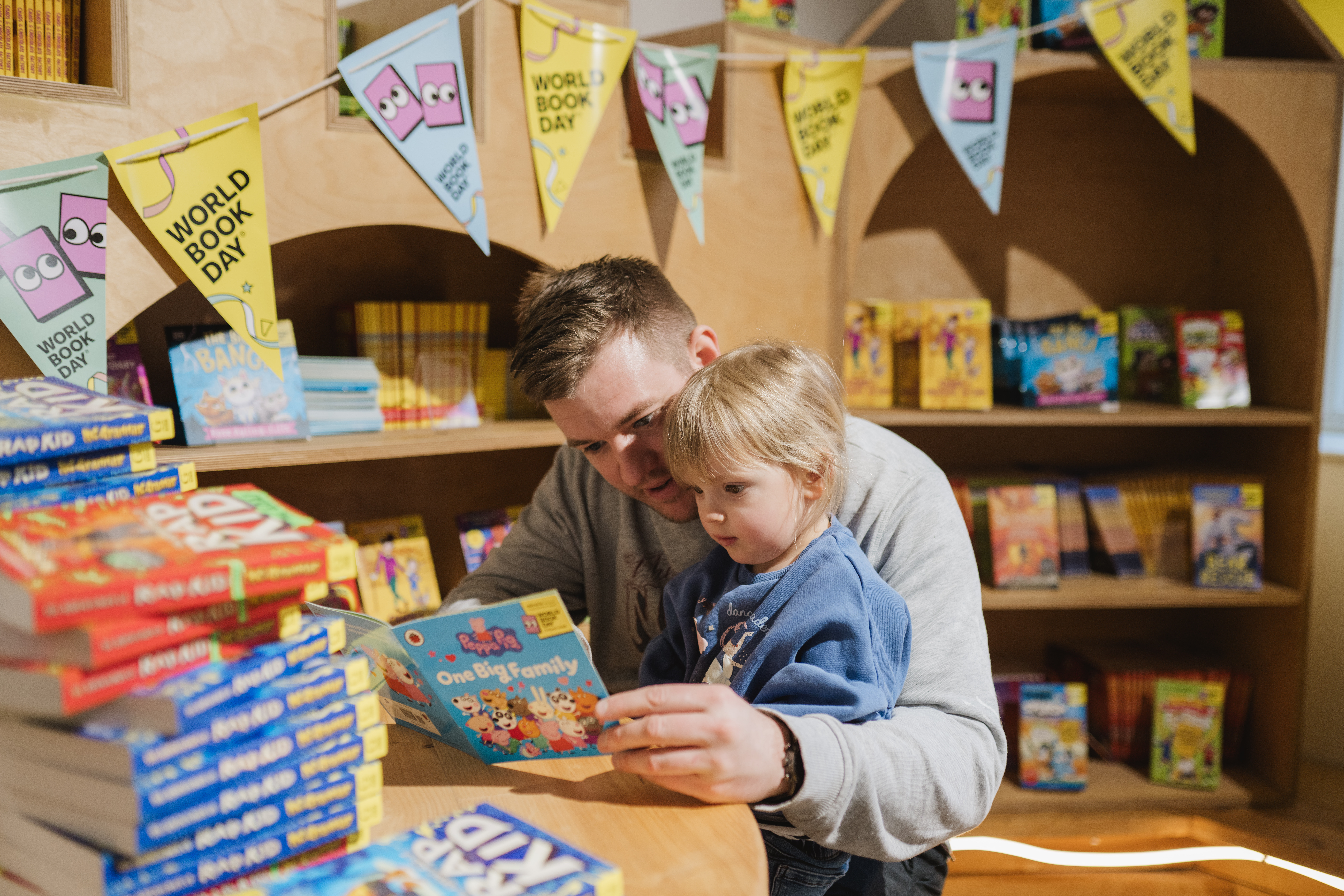 A man and a young child read a book together in a bookshop decorated with World Book Day banners, surrounded by colourful children's books.