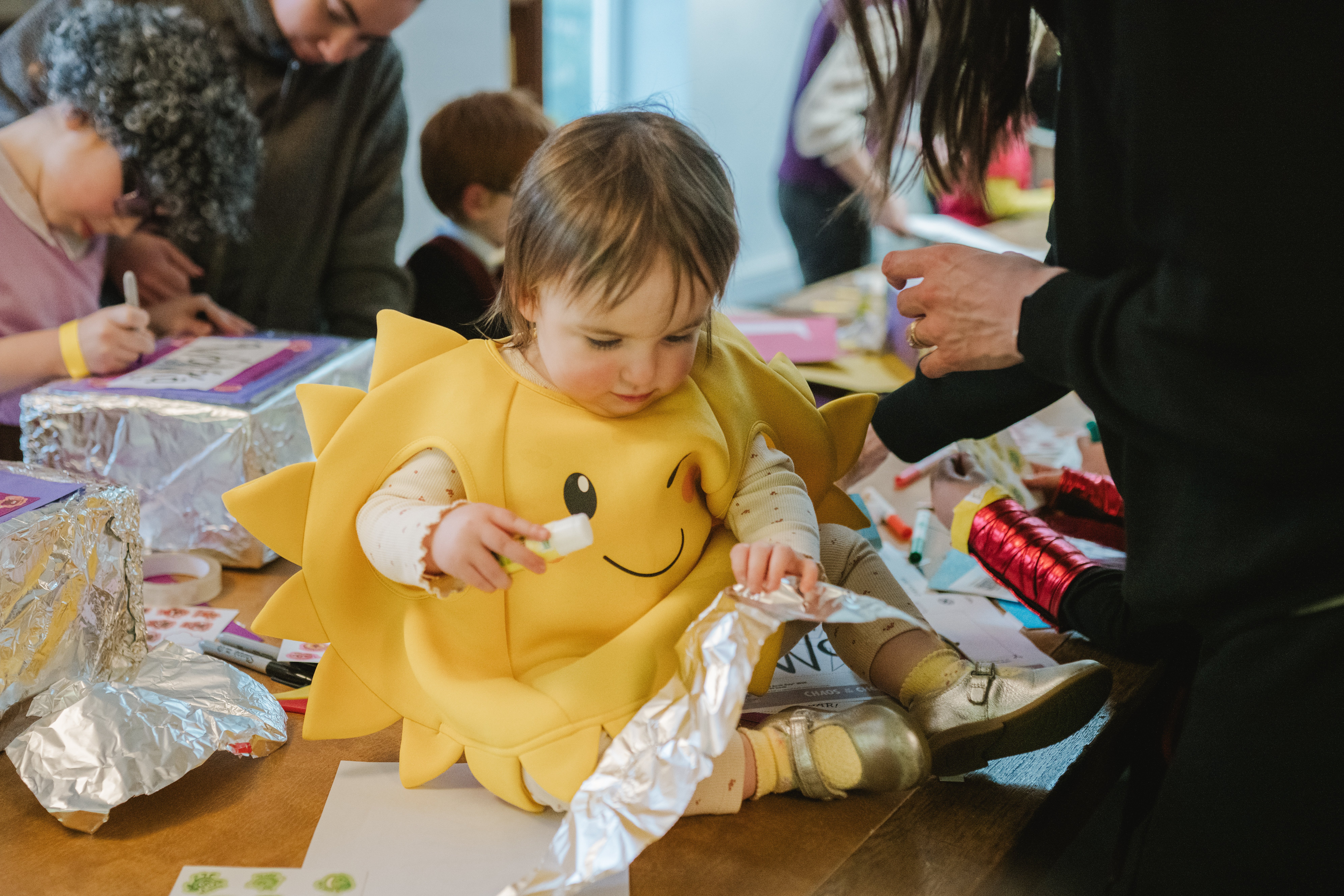 A toddler in a sun costume sits at a table with crafts, surrounded by foil and papers, while adults and children work on activities in the background.