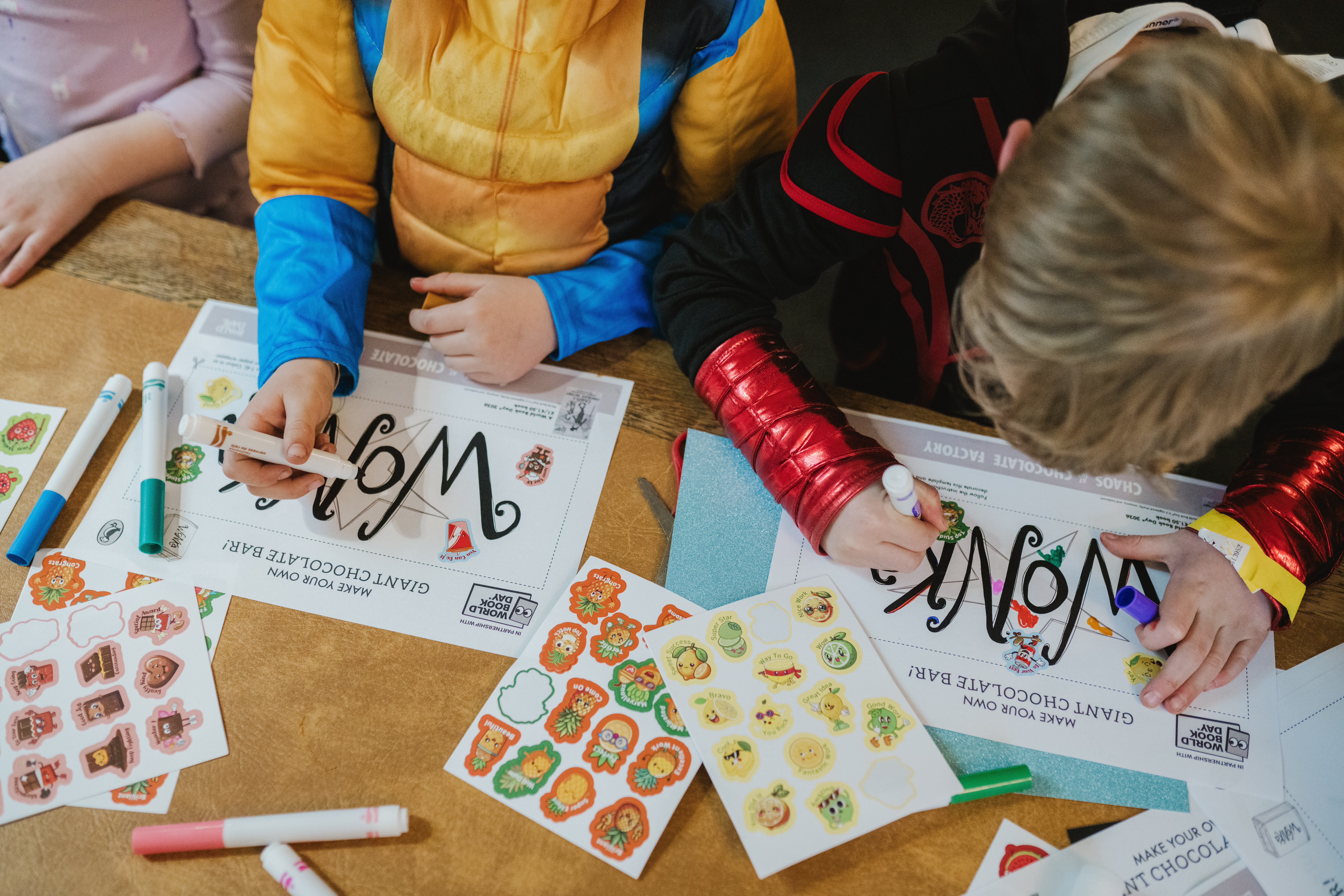 Children in colourful costumes decorate chocolate wrappers with markers and stickers at a craft table.