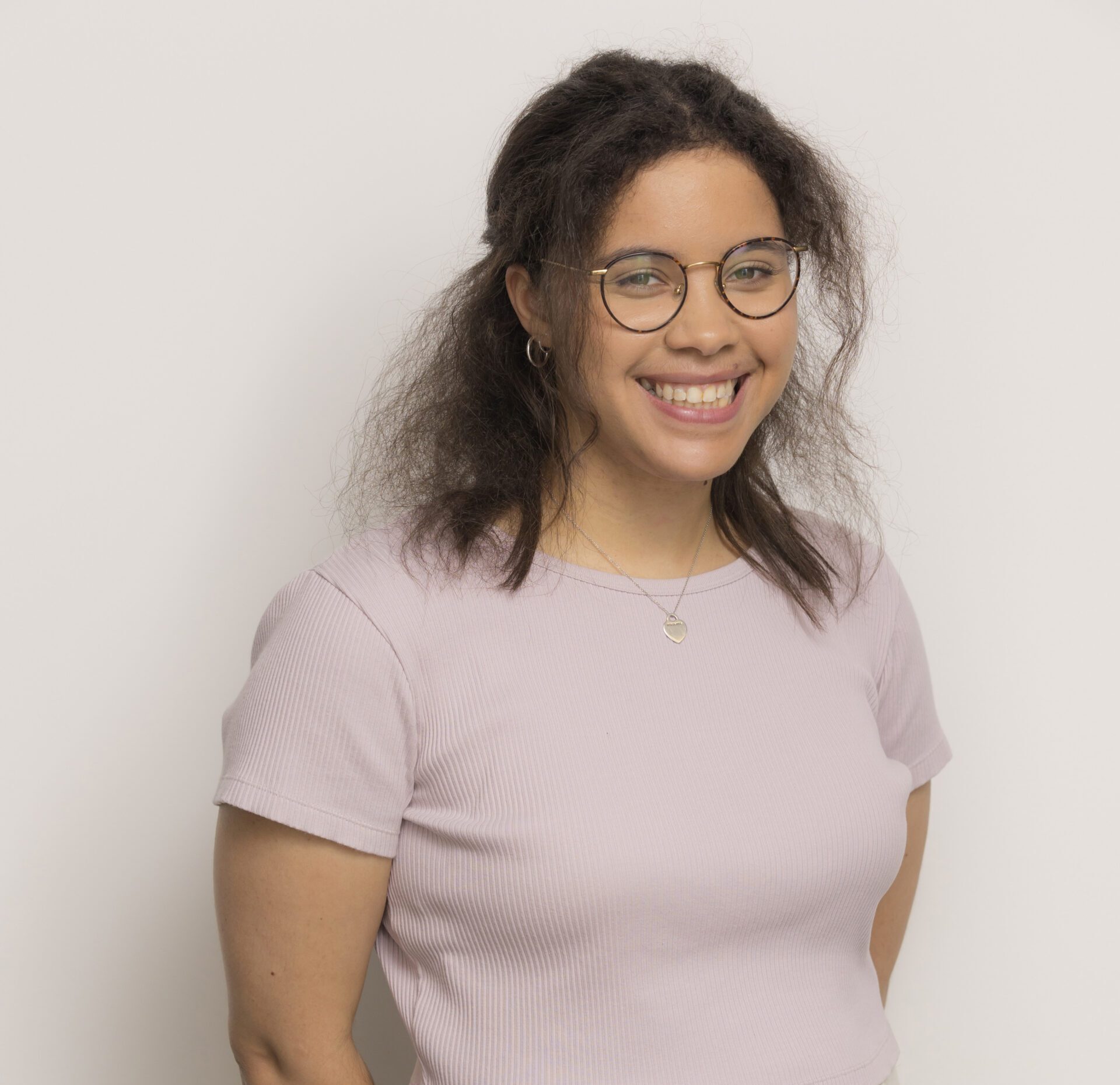 A smiling woman with glasses stands against a plain background, wearing a light purple top and cream trousers.