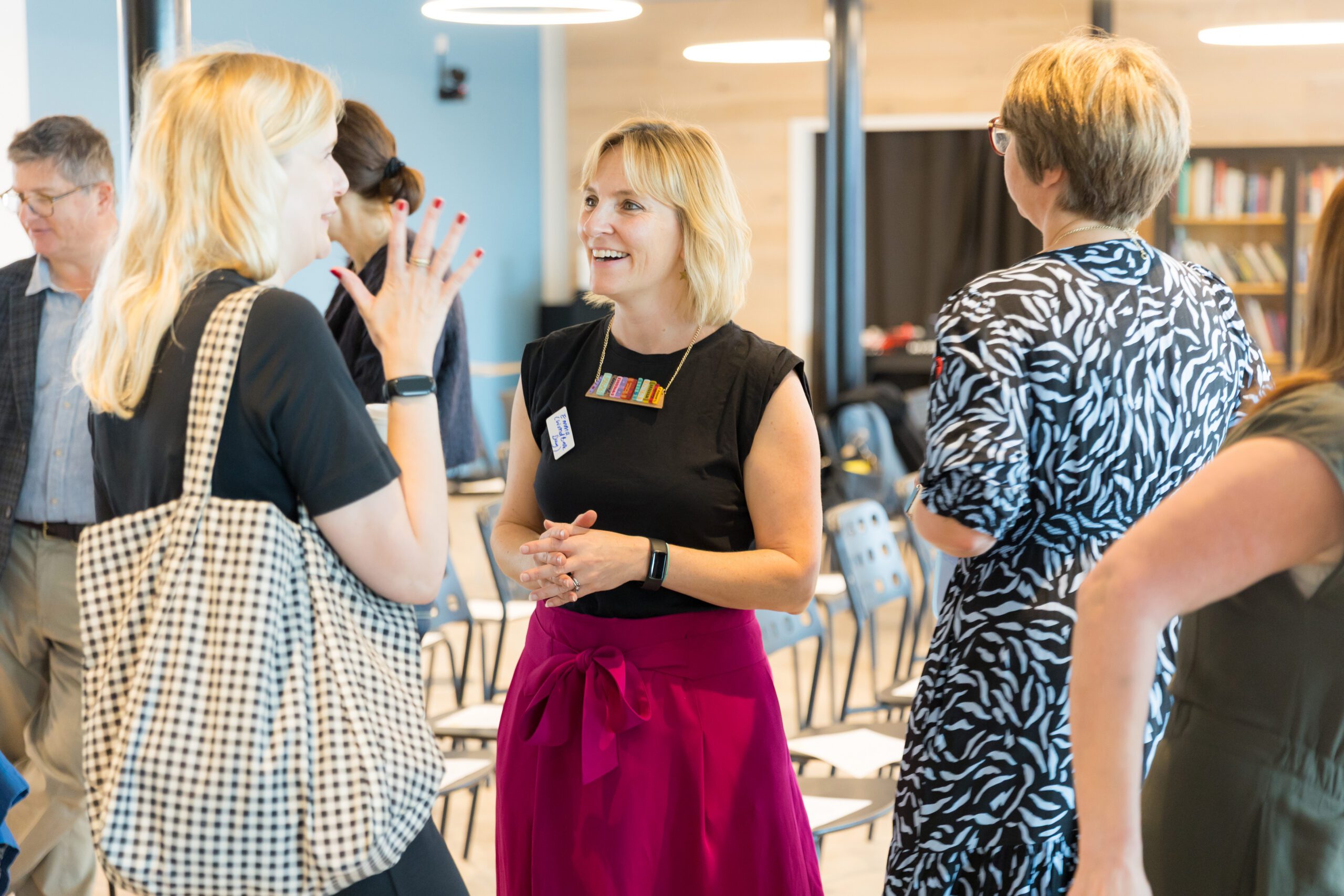 A group of people engaged in conversation at an indoor event, with a woman in the centre smiling and wearing a black top and pink skirt.